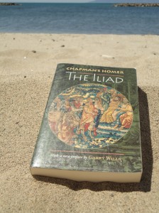 With sea in the background, on the sand of a beach sits Chapman’s Homer: The Iliad, the cover featuring a 16th-century Flemish tapestry (framed by a circle), Scene from Roman History, showing a number of men, the one in the middle armored; a horse is in the foreground